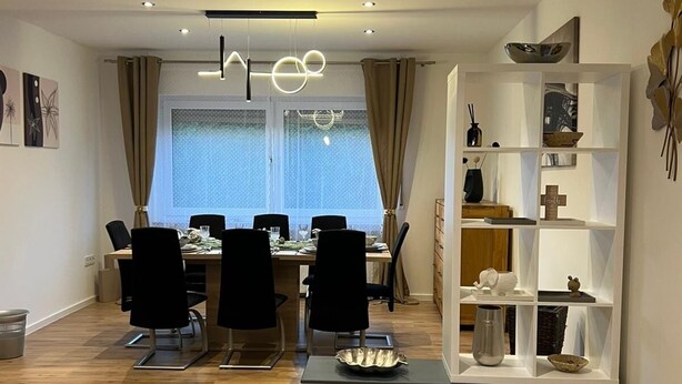 Un comedor moderno con suelo de madera, una mesa de comedor con sillas, una araña y una estantería decorativa con diversos objetos.