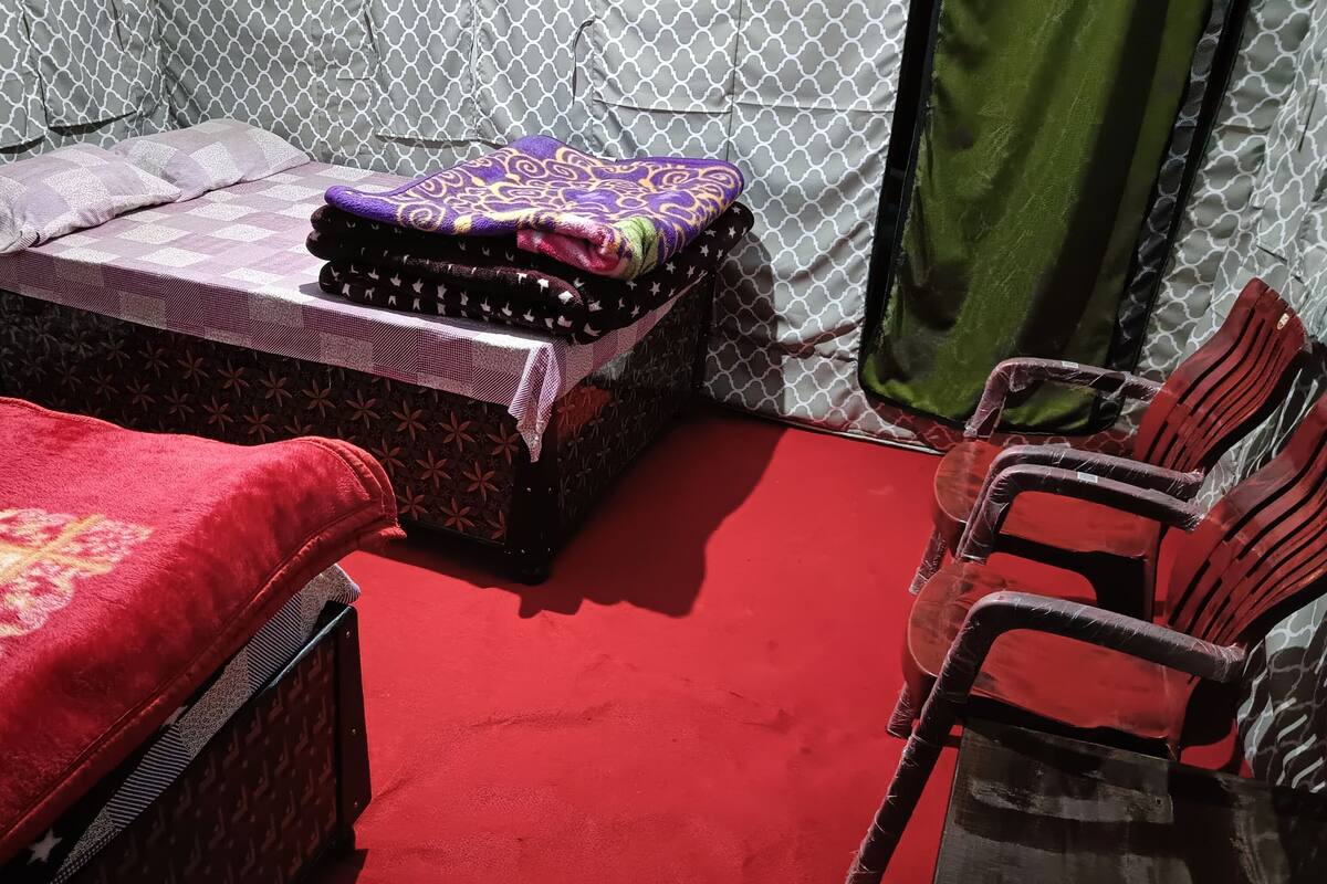 Une pièce avec une moquette rouge, un lit recouvert d’une couvre-lit à motifs, une petite table sur laquelle est posée une couverture violette, et un rideau vert.