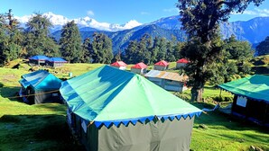 Ein Campingplatz mit mehreren Zelten, manche mit blauen und grünen Planen, vor einer Kulisse aus schneebedeckten Bergen und dichtem Wald.