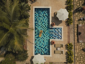 Vista aérea de una piscina rectangular con un diseño de baldosas a cuadros, rodeada de palmeras y mobiliario exterior con sombrillas.
