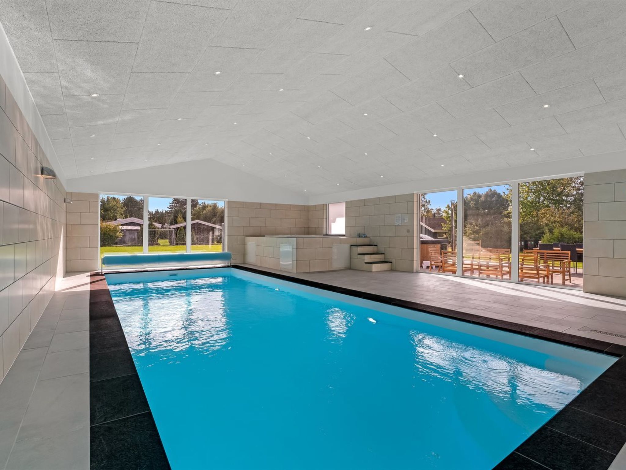 A modern indoor swimming pool with clear glass walls, a tiled floor, and a view of the outdoors.