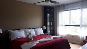 Une chambre d’hôtel avec un lit, un ventilateur de plafond, une fenêtre avec des rideaux et une table de chevet avec une lampe.