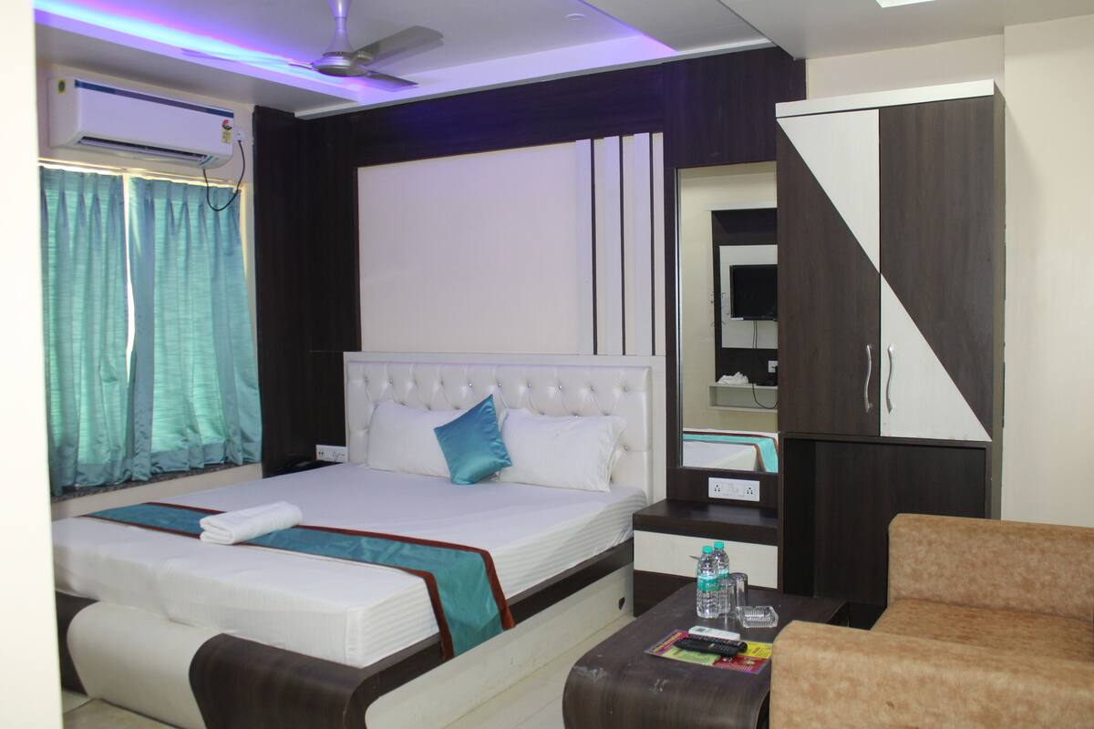 A hotel room with a large bed, a bedside table with a remote control, a water bottle, and a brown sofa.