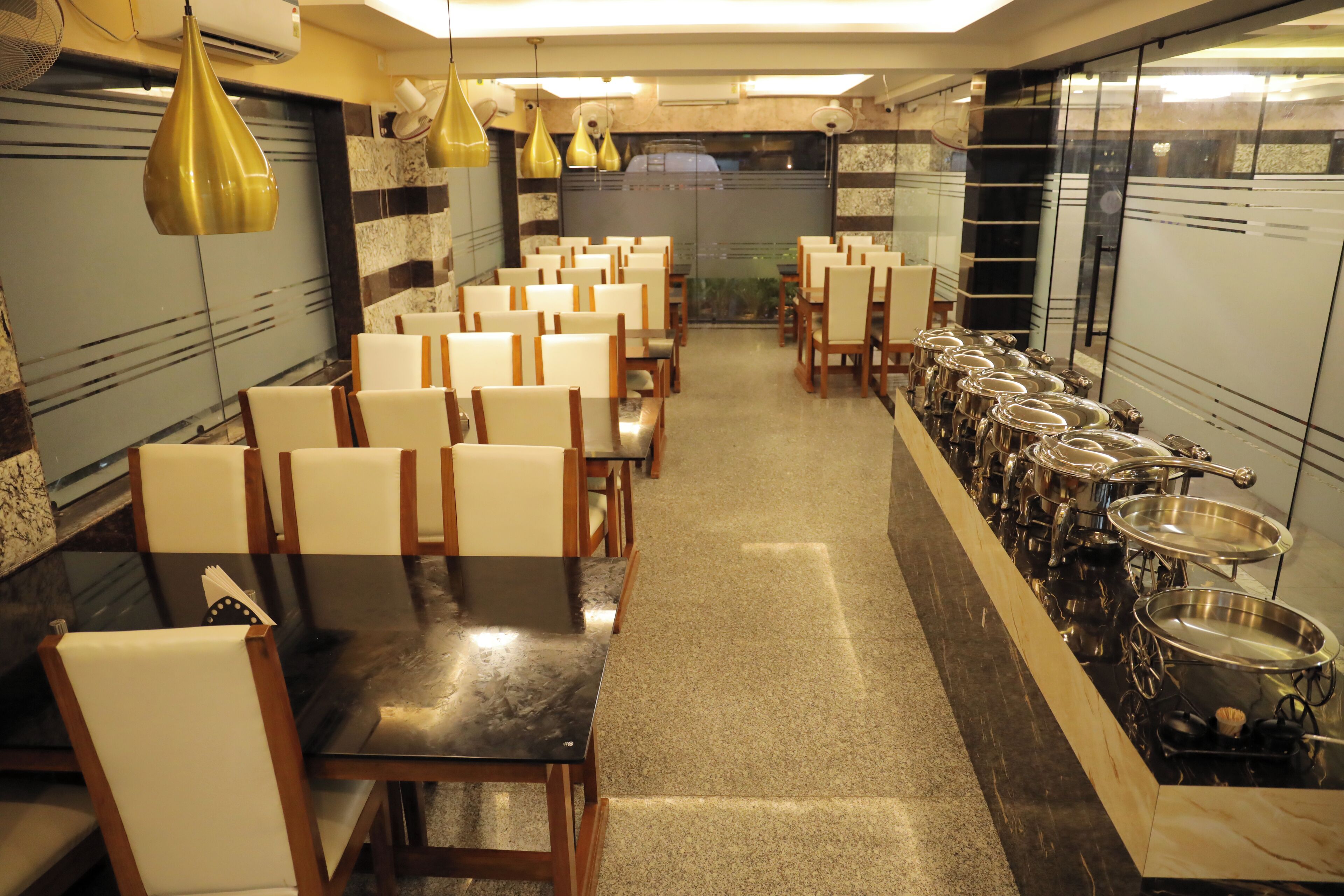 A restaurant with rows of tables and chairs, a long counter with cooking equipment, and hanging gold pendant lights.