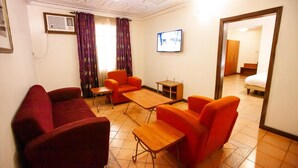 A hotel room with a red sofa, two orange armchairs, a small wooden table, and a TV mounted on the wall. - Genesis Place  (Port Harcourt)