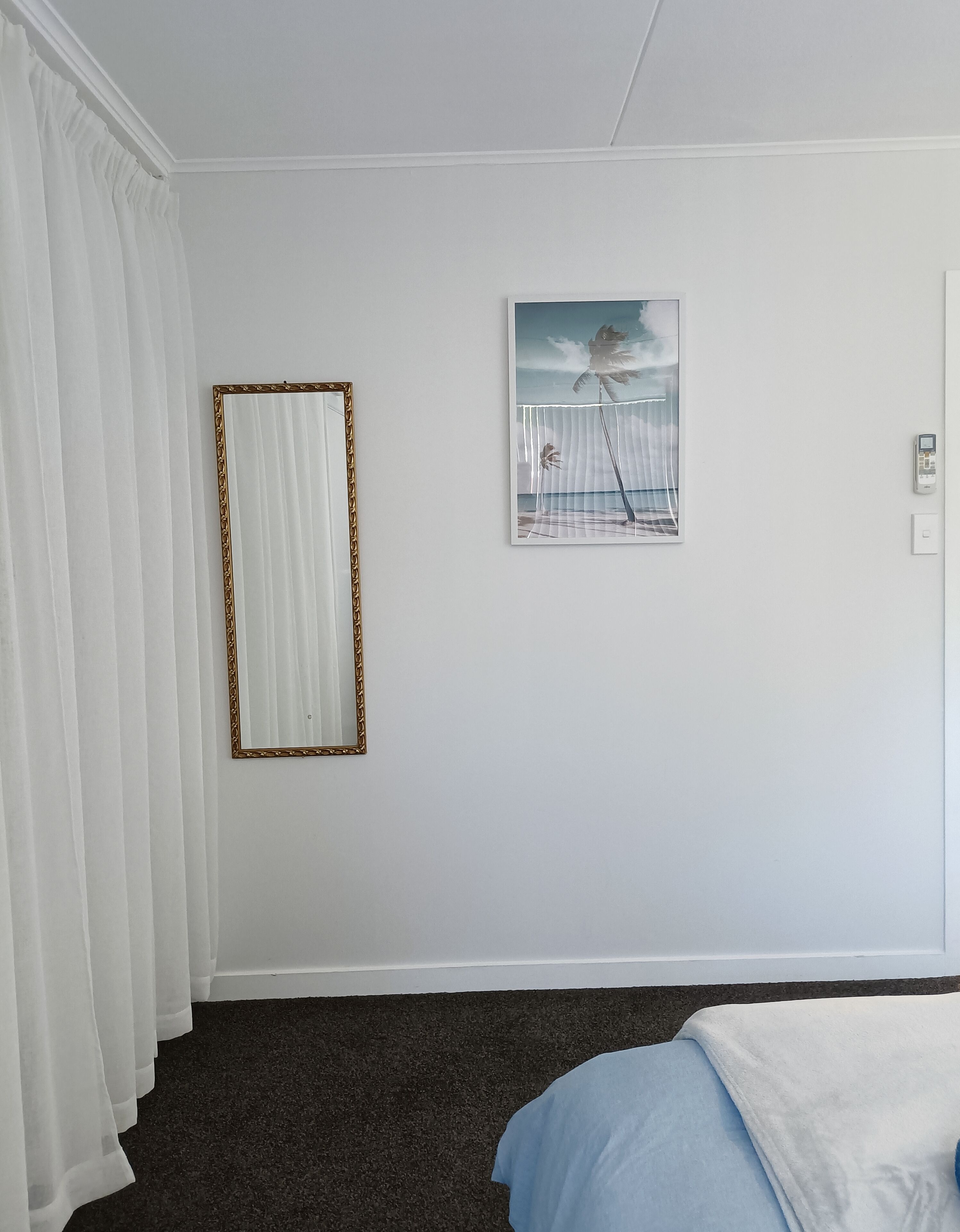 Une chambre d’hôtel avec un grand miroir, un tableau représentant une scène de plage, et un lit recouvert d’une couverture bleue.