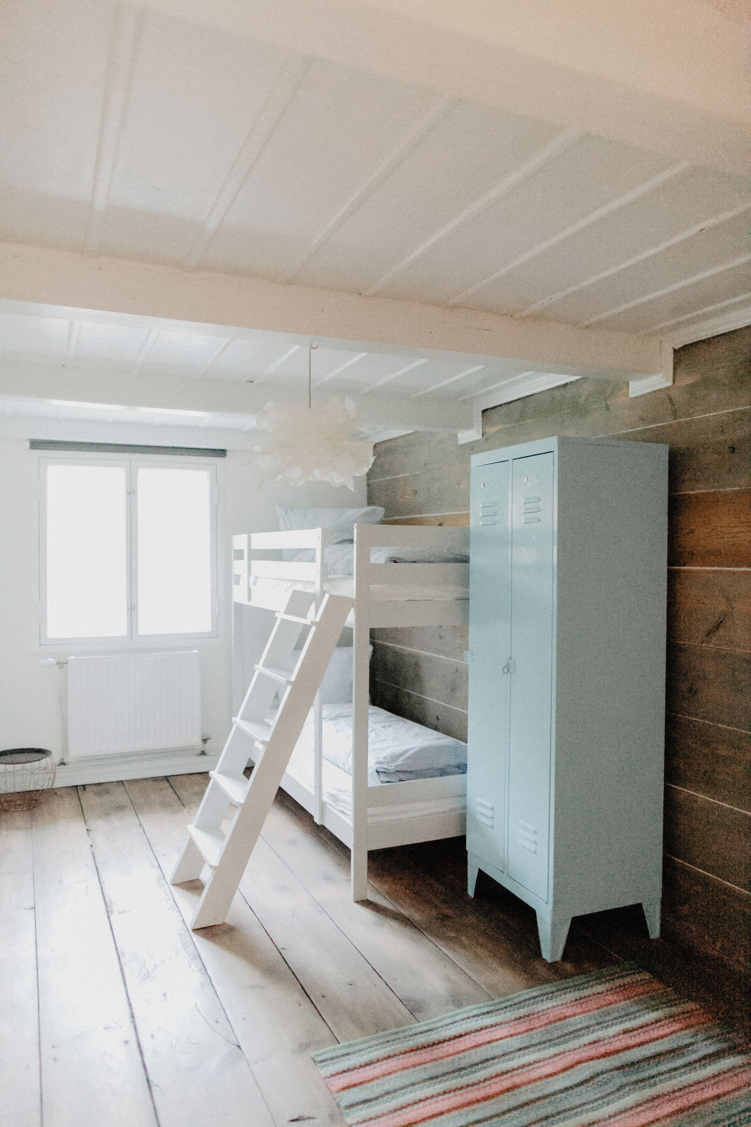 Una cama litera con escalera, un armario y una ventana en una habitación con suelo de madera y radiador.