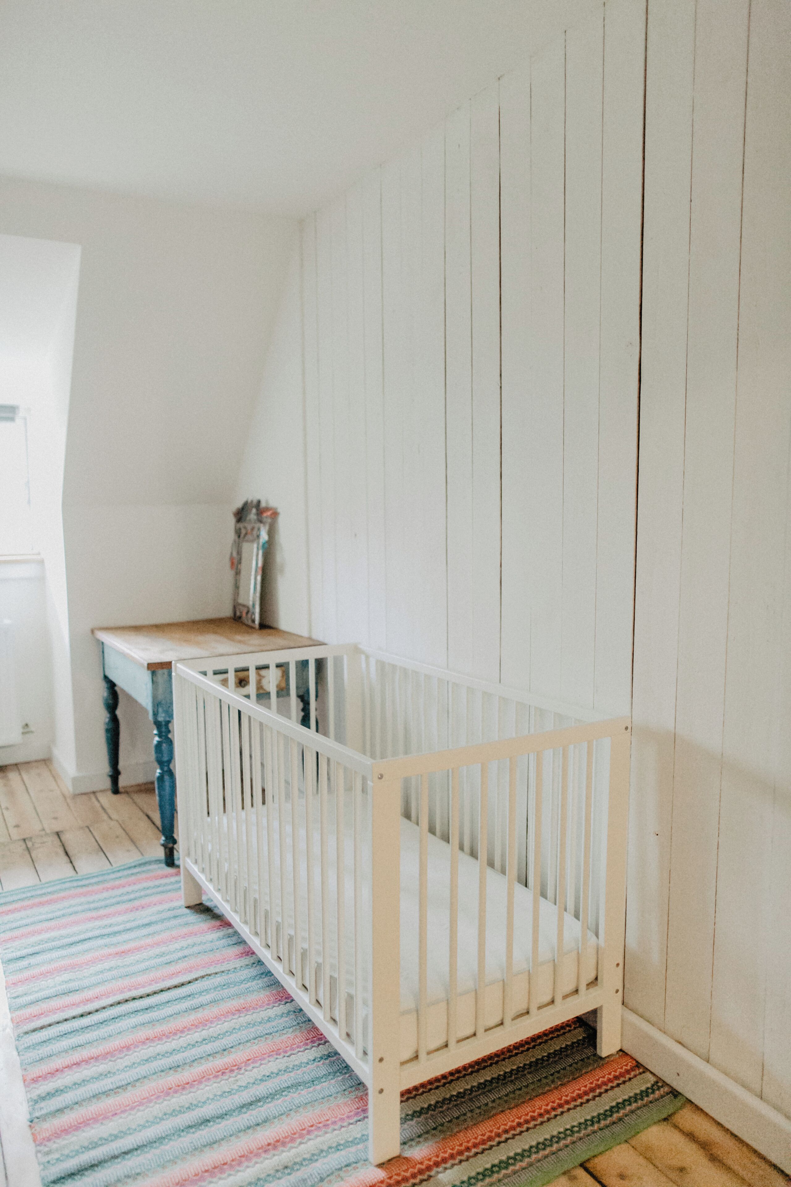 A room with a white crib, a wooden table, and a colorful striped rug.
