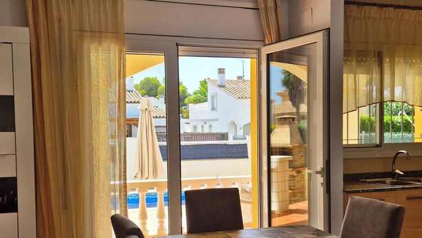 Un comedor con mesa y sillas, vistas a una piscina y edificios, y una cocina con lavamanos y armarios.