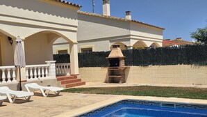 Una piscina con solárium empedrado, una parrilla y un área de estar techada con sombrillas.