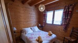 A wooden cabin bedroom with a bed, a nightstand, a chair, and a window with curtains. - Spacious chalet for 10 near slopes with private parking (Le Grand-Bornand)