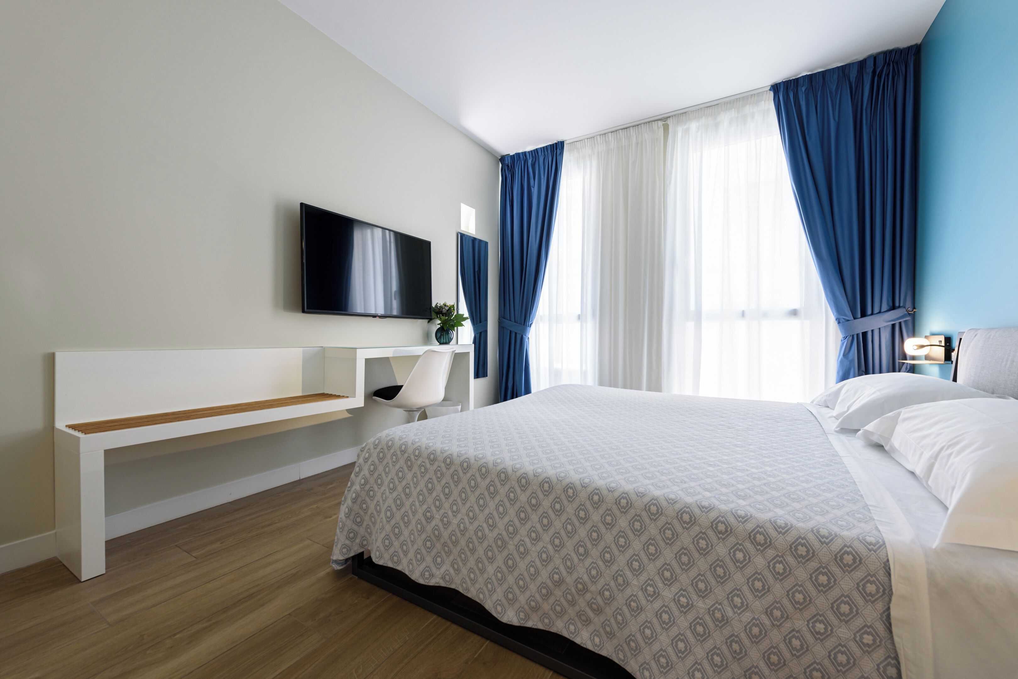 Habitación de hotel con cama, televisión montada en la pared, escritorio con silla y ventana con cortinas azules.