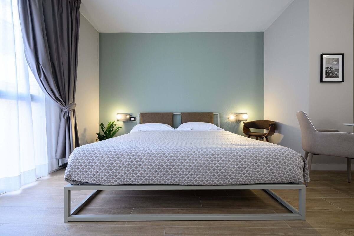 A modern hotel room with a bed, two bedside lamps, a chair, and a plant.