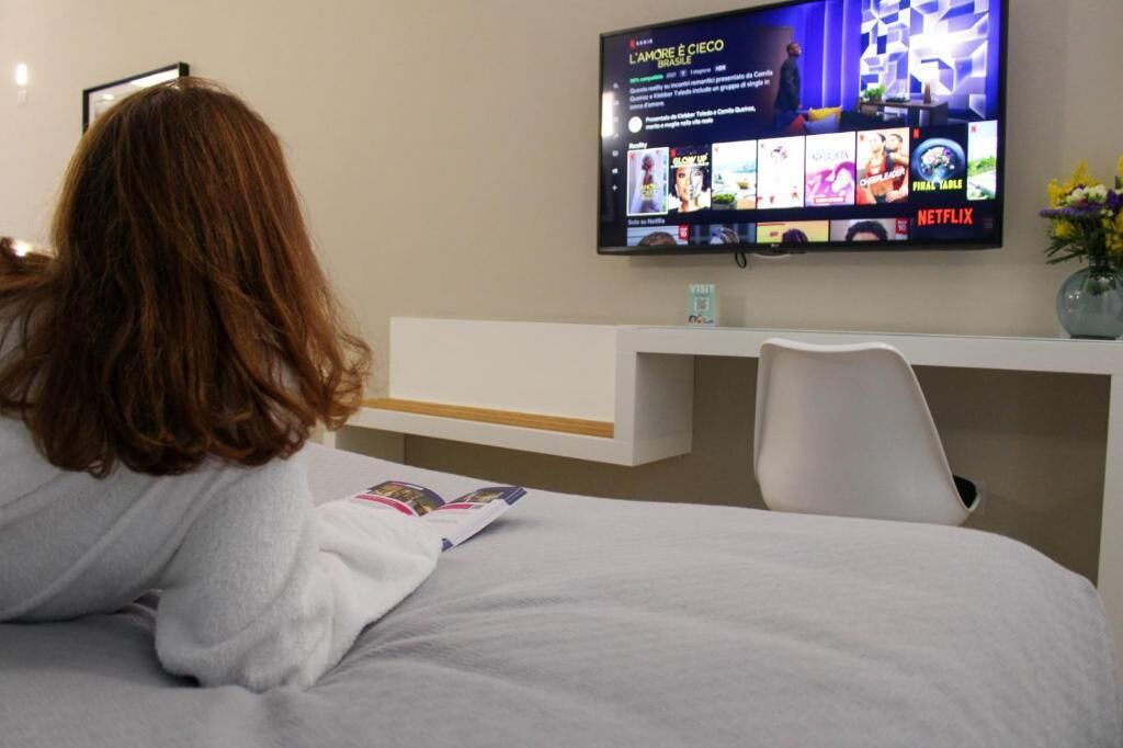 Een hotelkamer met een bed, een aan de muur bevestigde televisie met verschillende zenders, een wit bureau en een stoel.