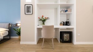 Una habitación pequeña con una cama, un escritorio con silla, un estante con objetos y un cuadro en la pared.
