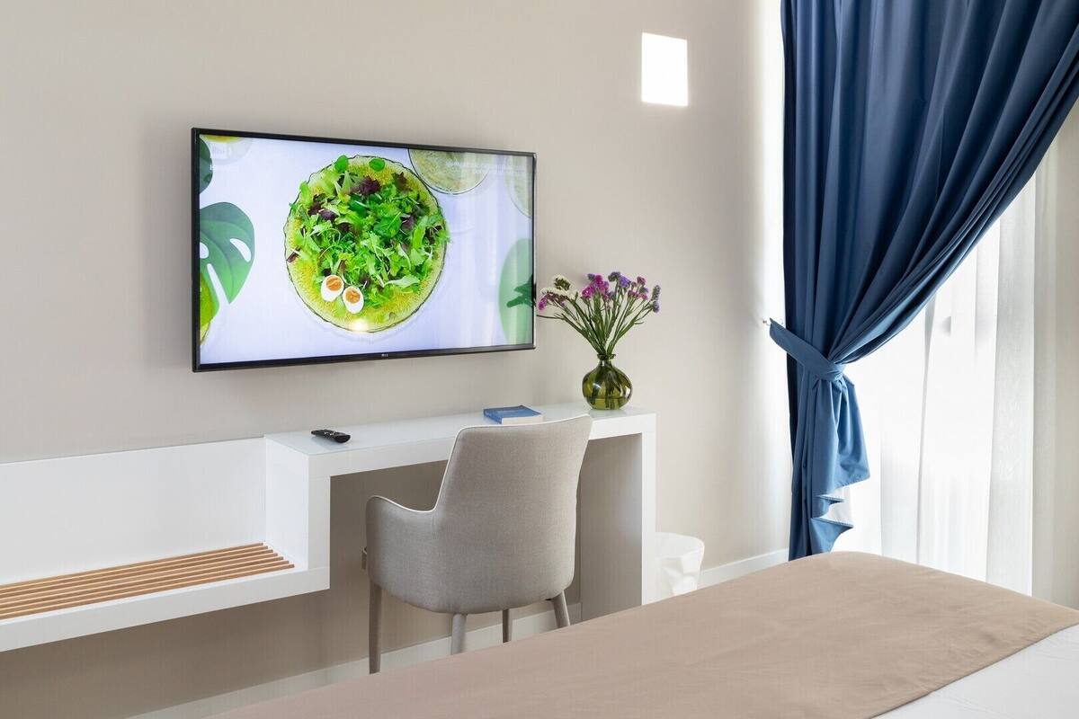 A modern hotel room with a white desk, a grey chair, a flat-screen TV mounted above the desk, a vase with flowers, and a window with blue curtains.