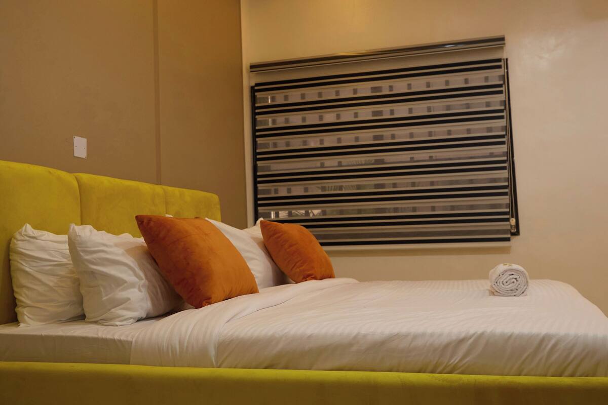 A bed with white and orange pillows, a yellow headboard, and a rolled towel on the bed.
