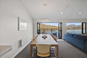 A modern dining area with a wooden table and chairs, a blue sofa, and a view of a lake and mountains.