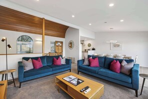 A modern living room with two blue sofas, a wooden coffee table, and a dining area with white chairs.