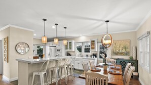 A modern kitchen with white cabinetry, a central island with bar stools, pendant lighting, and a dining area with a wooden table and chairs. - Lovely home with backyard oasis & great WiFi - walk to La Jolla Village, dogs OK (La Jolla)
