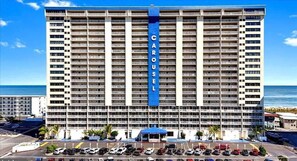 Um hotel com vários andares, com uma placa grande que diz 'CAROUSEL', um estacionamento e vista para o oceano.