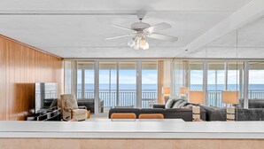 A spacious living area with a large window overlooking the sea, a ceiling fan, and a modern interior design. - Direct Oceanfront Condo with the Best Amenities (Ocean City)