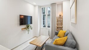 Ein modernes Wohnzimmer mit einem grauen Sofa, einem Holztisch, einem an der Wand montierten Fernseher und einem Fenster mit Vorhängen.