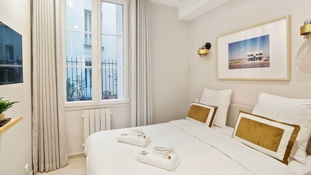 Ein Hotelzimmer mit einem großen Bett, einem Fenster mit Vorhängen, einem gerahmten Bild an der Wand und einem Fernseher links.