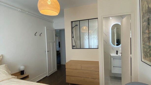 A modern bedroom with a large bed, a wooden dresser, a vanity with a mirror, and a hanging light fixture. - Maison Auguste (Paris)