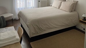 A bedroom with a bed, a chair, a desk, a radiator, and a window with curtains. - Maison Auguste (Paris)
