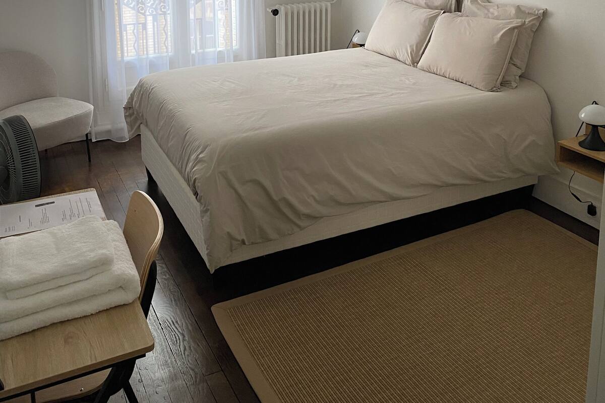 A bedroom with a bed, a chair, a desk, a radiator, and a window with curtains.
