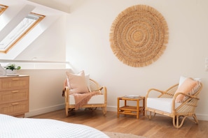 A room with two wicker chairs, a small table, a rattan wall hanging, and a wooden dresser.