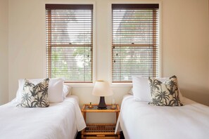 A room with two beds, each with a white bedspread and a pillow with a palm leaf pattern. There is a nightstand with a lamp and a glass of water between the beds. - "Treetop Retreat" – Ocean Views & minutes to Beach (MacMasters Beach)