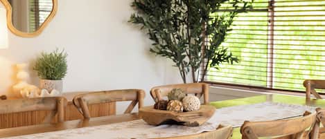 Ein Holztisch mit einer blumigen Tischläufer, umgeben von Holzstühlen. An der Wand hängt ein großer Spiegel, und im Hintergrund ist eine Topfpflanze zu sehen.