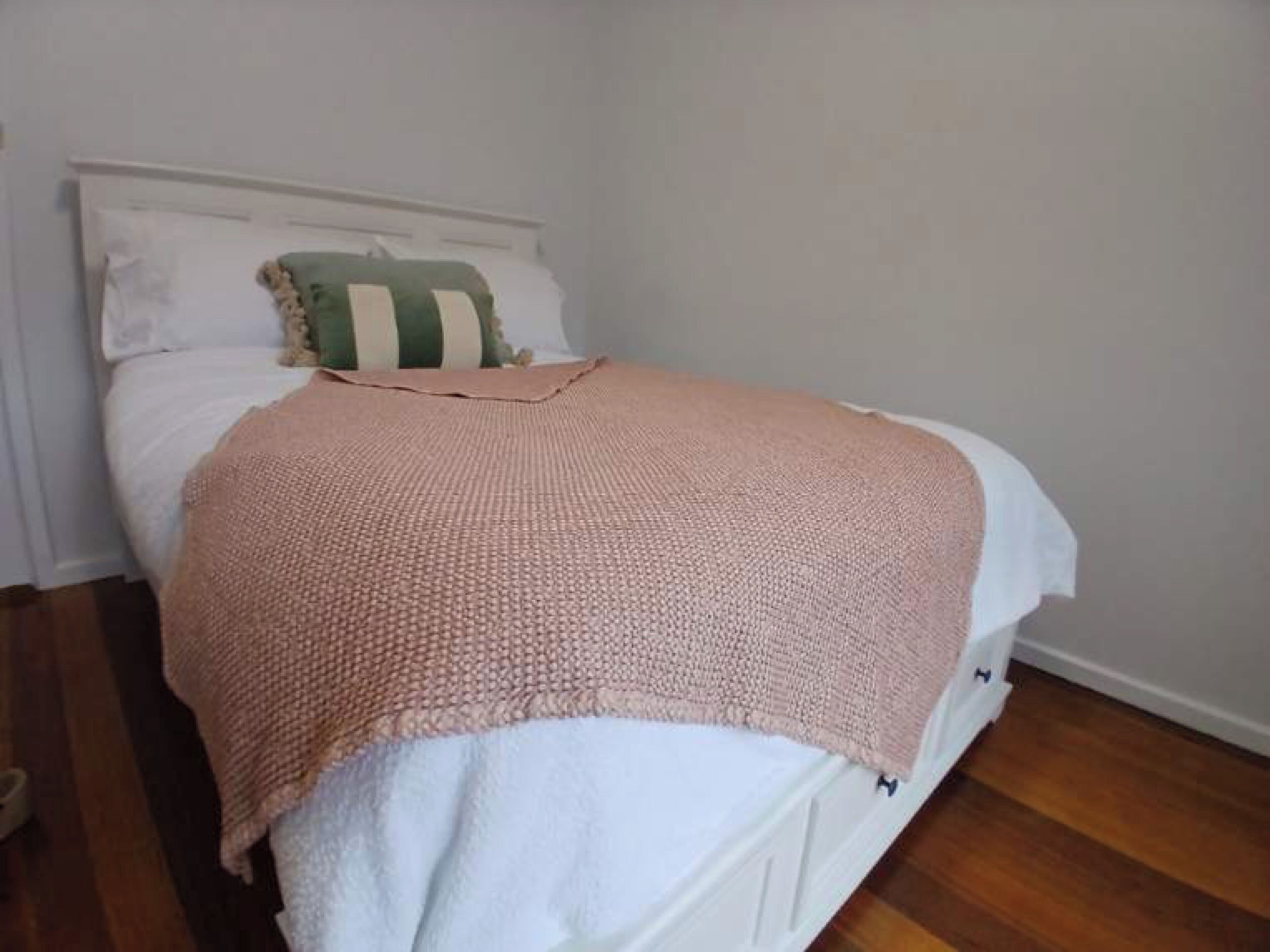 Ein ordentlich bezogenes Bett mit einer rosa gestrickten Decke, weißen Kissen und einem grün-weißen Streifenmuster-Kissen.