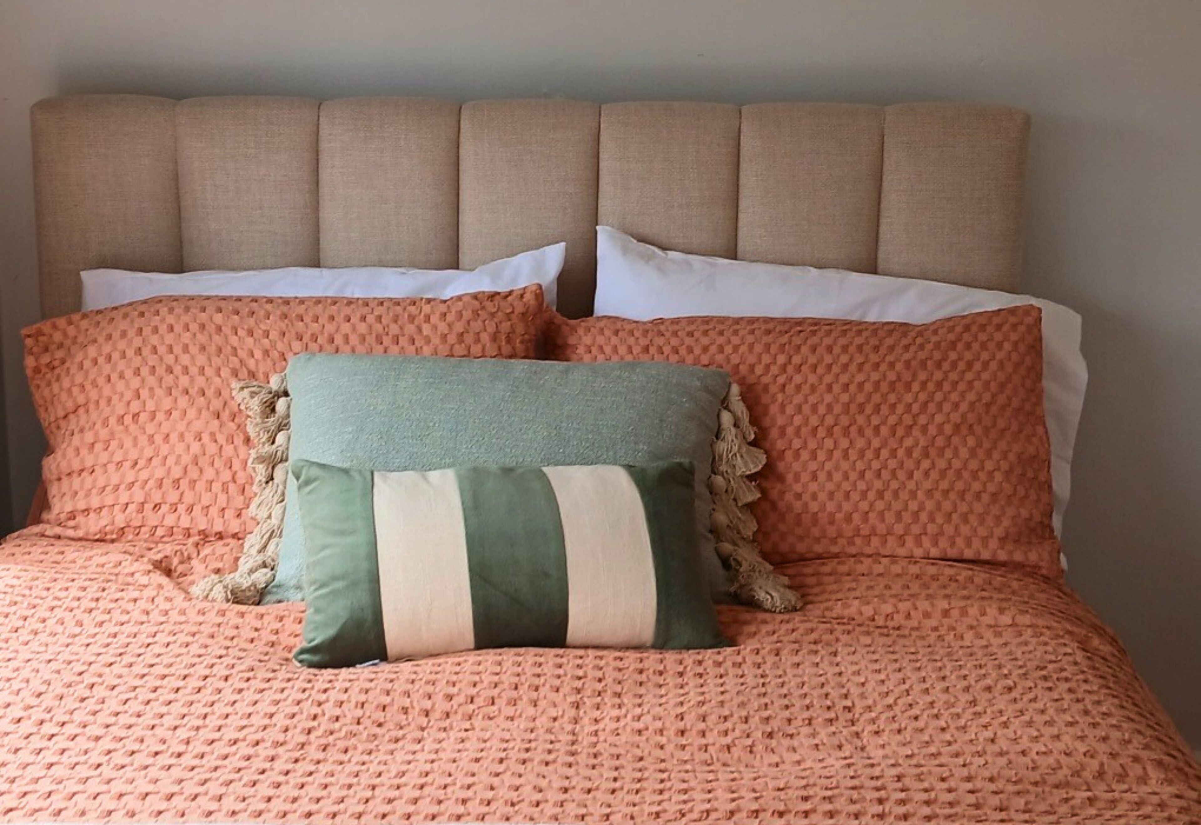 En säng med en beige stoppad sänggavel, två kuddar och ett persikofärgat, strukturerat överkast.