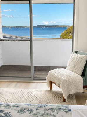 A room with a view of the sea, a sliding glass door, a white chair, a patterned rug, and a bed with a floral bedspread.