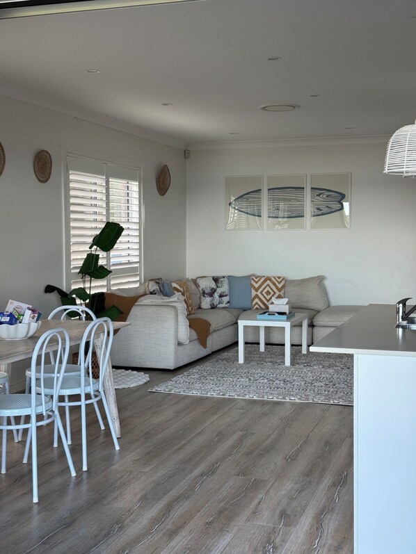 Ein modernes Wohnzimmer mit grauem Sofa, weißen Sesseln, einem kleinen weißen Tisch und einer Küchenzeile mit weißer Arbeitsplatte und Spüle.