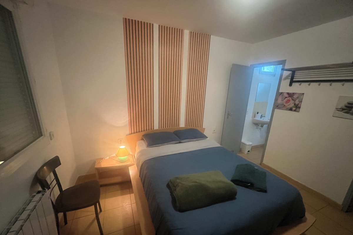 Une chambre à coucher comprenant un lit, une chaise, une table de chevet avec une lampe, une fenêtre et une porte donnant sur une salle de bain.