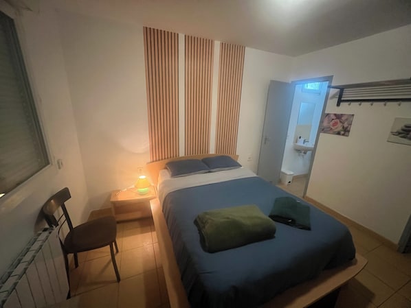 A bedroom with a bed, a chair, a nightstand with a lamp, a window, and a door leading to a bathroom. - Le Manteau Bleu (Béziers)