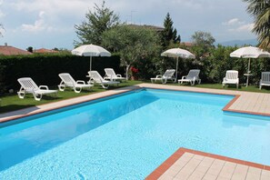 A swimming pool with white lounge chairs and umbrellas, surrounded by greenery and a tiled deck. - Ferienwohnung mit Terrasse und Pool (Lazise)