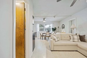 A modern living room with a beige sofa, a dining area with a table and chairs, a ceiling fan, and a window with blinds.