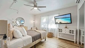 Quarto com cama, mesa de cabeceira, espelho, televisão e vista para a praia.