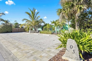 A paved driveway leading to a building with a number sign '05' in a garden with palm trees and shrubs.