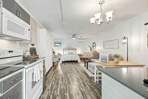A modern kitchen with a microwave, oven, and a coffee machine. There is a dining area with a table and chairs. The room has a bed with a white comforter and a ceiling fan.