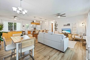Ein modernes Wohnzimmer mit Sofa, Esstisch und Stühlen, eine Küche mit Holzausbau und einem Fernseher, der eine Strandansicht zeigt.
