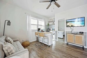A modern living room with a sofa, a dining area with a small table and chairs, a kitchen with a TV, and a ceiling fan. - Sandpiper Studio | Across From Beach! (Satellite Beach)