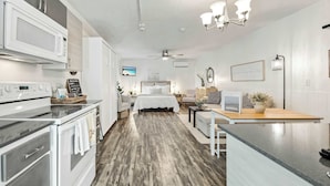 A modern kitchen with a microwave, oven, and a coffee machine. There is a dining area with a table and chairs. The room has a bed with a white comforter and a ceiling fan.