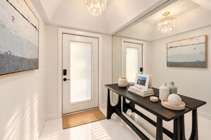 Un couloir avec une porte, un miroir, une console ornée d’objets décoratifs et un lustre.