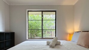 Une chambre avec une grande fenêtre, un lit recouvert de draps blancs et une lampe de chevet.